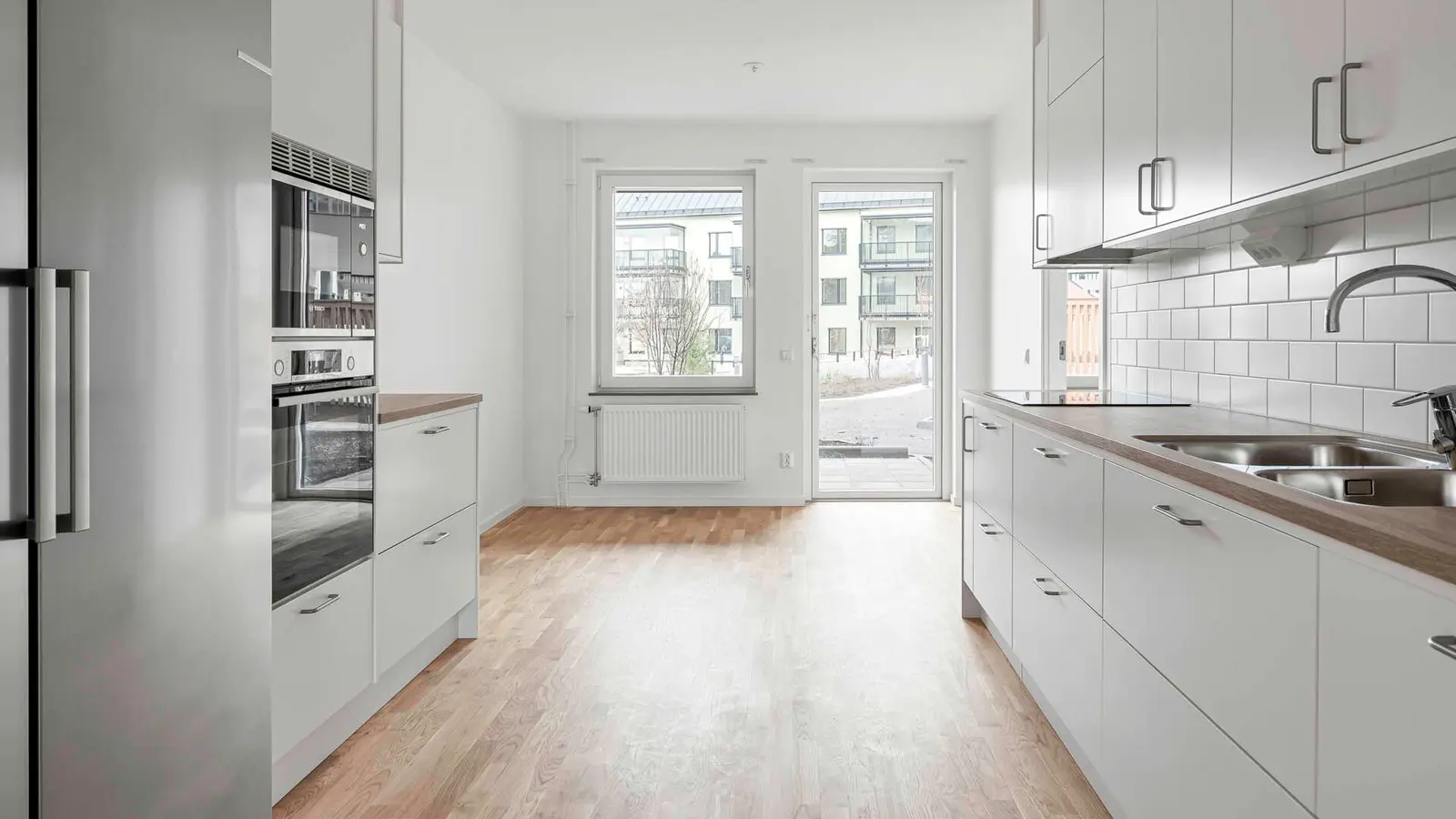 Modernt kök med vita skåp, inbyggd ugn och mikrovågsugn, träparkettgolv samt utsikt mot en innergård genom ett stort fönster och altandörr